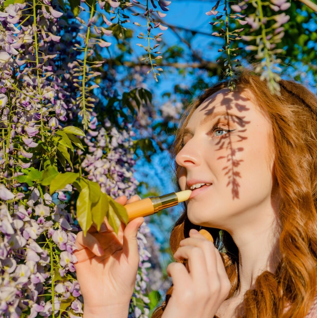 “Woman applying The Bee Fairy natural beeswax lip balm outdoors, surrounded by greenery and sunshine — promoting eco-friendly handmade skincare.”